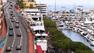 Carros de formula 1 alinhados em mônaco com iates e prédios ao fundo.